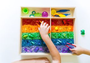 Sensory box with rainbow rice inside. Children's hands touch the texture and pour. Tool for sorting tongs and spoon. Montessori material baby