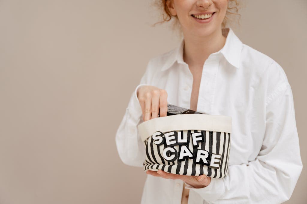 A smiling woman in a white shirt holding a striped bag labeled 'SELF CARE' indoors.