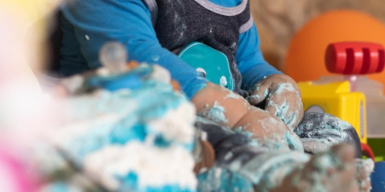Unrecognizable barefoot toddler in comfortable wear playing with light blue sugar cream for festive cake